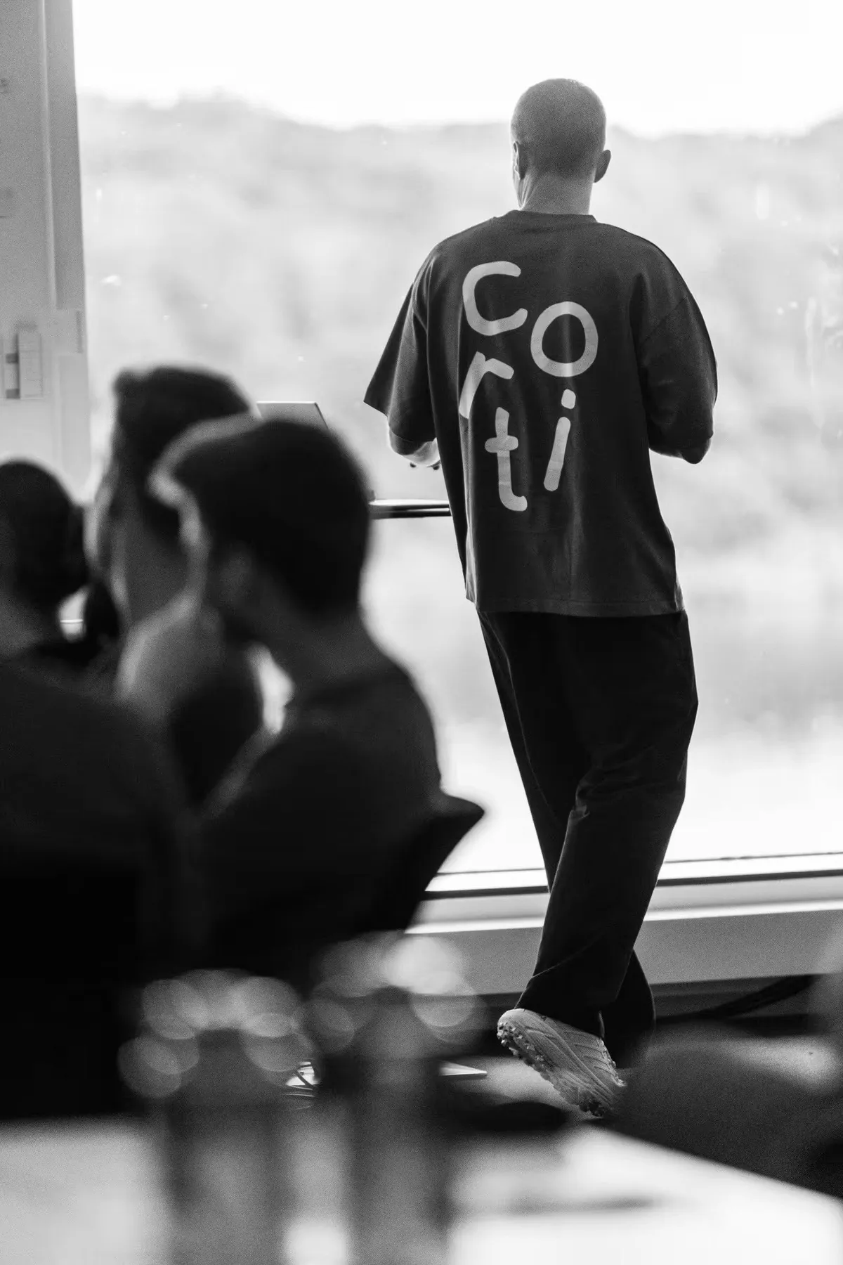A Cortian in a Corti t-shirt standing by a window in front of colleagues